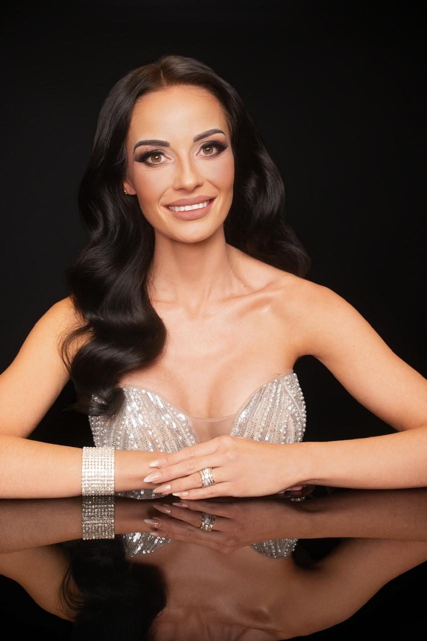 Andrea Raaths Duarte in a strapless silver beaded gown, hands folded on a black reflective table, studio portrait against black